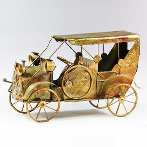 Puede incluir: Modelo de coche antiguo de latón con detalles intrincados. El modelo presenta un techo, faros y ruedas de radios. El latón tiene un aspecto envejecido, lo que aumenta su encanto antiguo. Artículo decorativo.