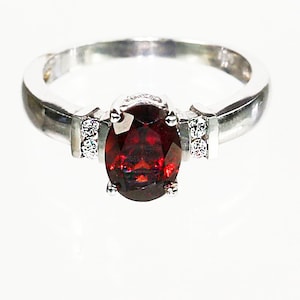 May include: A silver ring featuring a red oval gemstone with two smaller clear gemstones on either side.