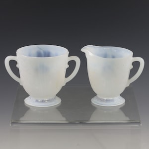 May include: A vintage white glass sugar bowl with two handles and a matching creamer. Both pieces have a floral pattern and sit on a clear glass surface. The interior of the bowl and creamer have a light blue hue.