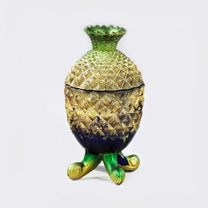 May include: A gold and green pineapple-shaped glass jar with a blue base and three green feet. The jar has a textured surface and a lid.