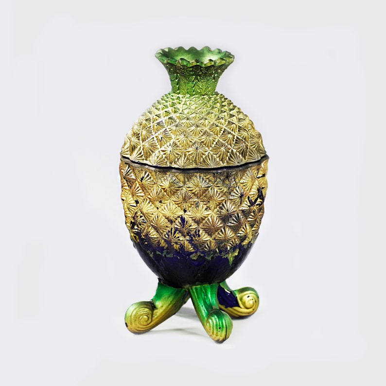 May include: A gold and green pineapple-shaped glass jar with a blue base and three green feet. The jar has a textured surface and a lid.