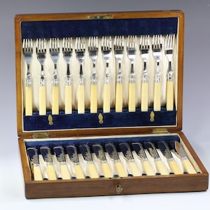 Puede incluir: Una caja de madera con un forro de terciopelo azul contiene un juego de 24 tenedores de plata con mangos de marfil. Los tenedores están dispuestos en dos filas de 12. La caja está abierta y los tenedores son visibles.