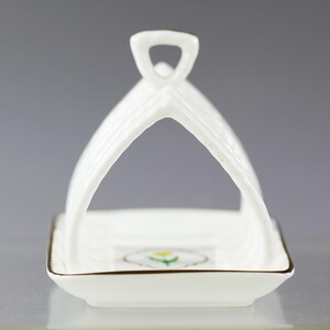 Puede incluir: Un tostador de porcelana blanca con asa. El soporte tiene una base cuadrada con ribete dorado y un dise&ntilde;o floral. El asa es arqueada y acanalada. El soporte est&aacute; dise&ntilde;ado para sostener las tostadas en posici&oacute;n vertical.