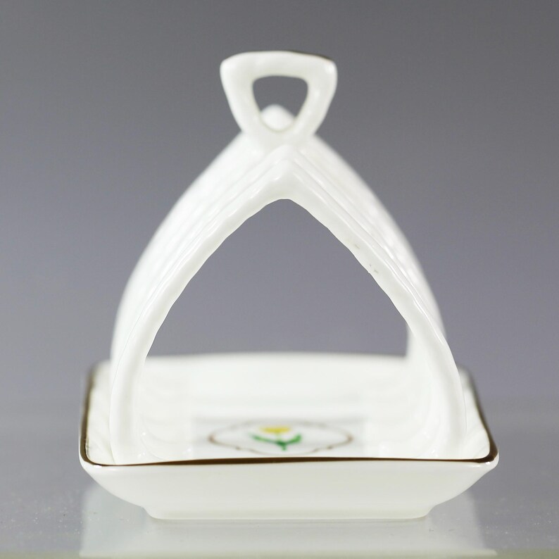 Puede incluir: Un tostador de porcelana blanca con asa. El soporte tiene una base cuadrada con ribete dorado y un dise&ntilde;o floral. El asa es arqueada y acanalada. El soporte est&aacute; dise&ntilde;ado para sostener las tostadas en posici&oacute;n vertical.