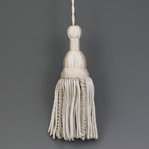 May include: A white tassel with a round top and a long, fringed bottom. The tassel is made of a woven material and has a delicate, elegant look.