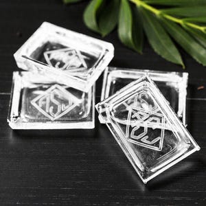 May include: Four clear glass coasters with a geometric design. Each coaster has a rectangular shape and features an etched monogram within a diamond shape. The coasters are arranged on a dark surface.