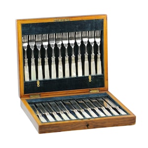 Puede incluir: Un juego de cubiertos de madera vintage con tapa con bisagras, que contiene tenedores y cuchillos. La cubertería presenta mangos plateados ornamentados y empuñaduras blancas nacaradas. El interior está forrado con terciopelo azul oscuro.