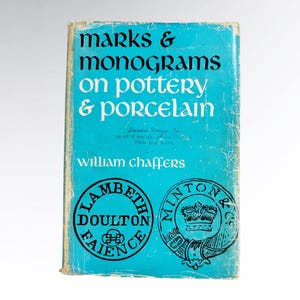 May include: A vintage book titled "Marks & Monograms on Pottery & Porcelain" by William Chaffers. The book has a turquoise cover with white and black text. The cover features two circular logos, one for Lambeth Doulton Faience and another for Minton & Co.