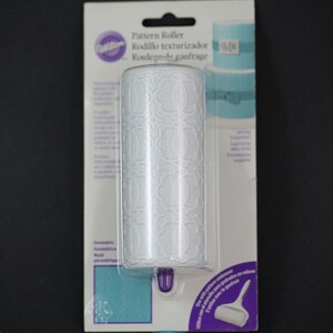 May include: White plastic rolling pin with a geometric pattern on the surface. The rolling pin is packaged on a white and purple card with the text "Pattern Roller" and "Wilton" on it.