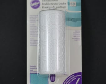 Puede incluir: Rodillo de plástico blanco con un patrón geométrico en la superficie. El rodillo está empaquetado en una tarjeta blanca y morada con el texto "Pattern Roller" y "Wilton" en ella.