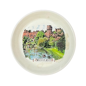 May include: A small, off-white ceramic plate featuring a colorful illustration of Warwick Castle. The image depicts the castle, a river, trees, and the text "Warwick Castle". The plate has a slightly worn appearance.