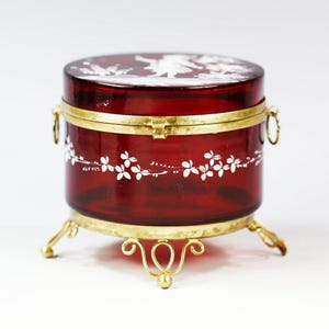 May include: A red glass trinket box with a white floral design and a gold metal hinged lid and base. The box is decorated with a white silhouette of a woman and flowers.