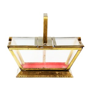 May include: A vintage-style, rectangular glass and brass box with a handle. The box has a red velvet interior and is constructed with clear glass panels and a gold-colored metal frame. The handle is also gold-colored.