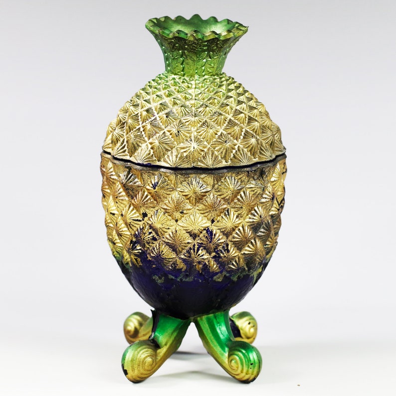 May include: A gold and green pineapple-shaped decorative jar with a blue base and three legs. The jar has a lid with a textured design.