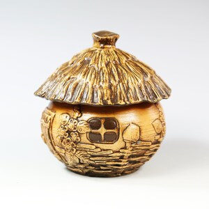 Op de afbeelding: Een keramische pot met een deksel in de vorm van een rieten dak. De pot is bruin met bruine accenten en heeft een gedetailleerd reliëf van een huisje met een raam, bloemen en een deur. Het deksel is donkerbruin en imiteert stro.