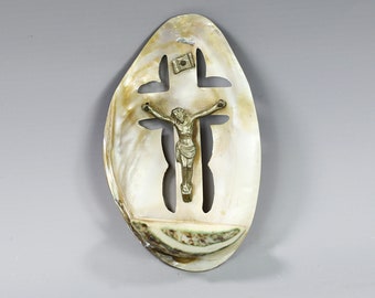 Peut inclure: Une coquille de nacre blanche avec une découpe en forme de croix et une petite figurine en métal de Jésus-Christ sur la croix. La coquille est de forme ovale et a une texture naturelle et organique.