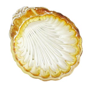 Puede incluir: Un plato decorativo en forma de concha con borde festoneado. El plato presenta un interior blanco con diseño acanalado y un borde marrón dorado. El borde tiene un aspecto texturizado y envejecido. El plato es probablemente de vidrio o material similar.