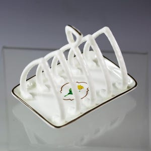 Puede incluir: Un tostador de porcelana blanca con base cuadrada y separadores arqueados. El tostador tiene un borde dorado y un dise&ntilde;o floral. El dise&ntilde;o presenta una flor amarilla con hojas verdes.