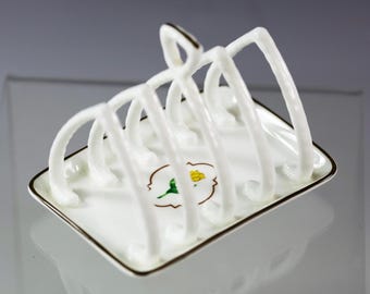 May include: A white porcelain toast rack with a square base and arched dividers. The rack has a gold trim and a floral design. The design features a yellow flower with green leaves. The rack is designed to hold toast.