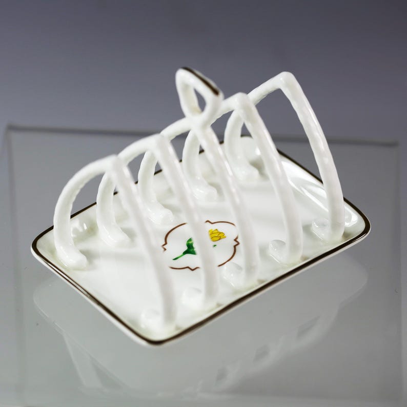Puede incluir: Un tostador de porcelana blanca con base cuadrada y separadores arqueados. El tostador tiene un borde dorado y un dise&ntilde;o floral. El dise&ntilde;o presenta una flor amarilla con hojas verdes.