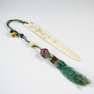 Puede incluir: Un marcapáginas de hueso blanco con un diseño floral y un borla. La borla está hecha de hilo azul y verde y tiene una cuenta rosa.