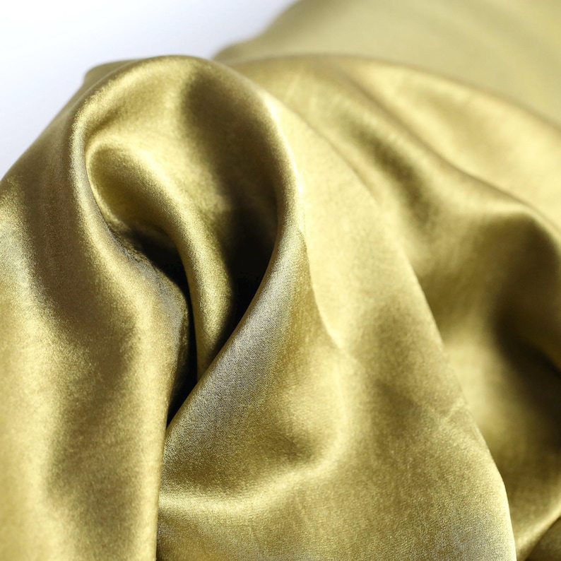 May include: Close-up of a flowing fabric in a golden-olive color. The material has a silky sheen, with soft folds and highlights. The fabric appears to be lightweight and drapes gracefully, suggesting it could be used for apparel or home decor.