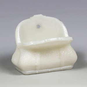 May include: A white, opaque, glass soap dish shaped like a purse with a textured surface. The dish has a small hole at the top for hanging.