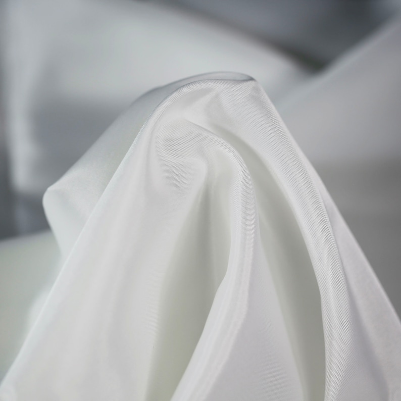May include: Close-up of a white fabric with soft folds and wrinkles. The fabric is smooth and silky, with a subtle sheen.