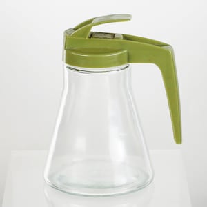 May include: A clear glass syrup dispenser with a green plastic lid and handle. The dispenser has a spout for pouring syrup.