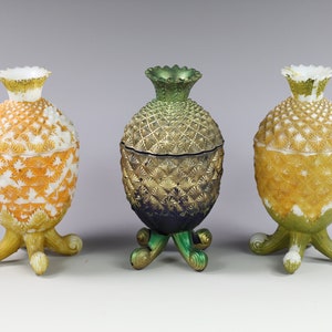 May include: Three decorative pineapple-shaped containers with lids. The containers are made of glass and have a white, yellow, and green color scheme. The containers are on a white background.