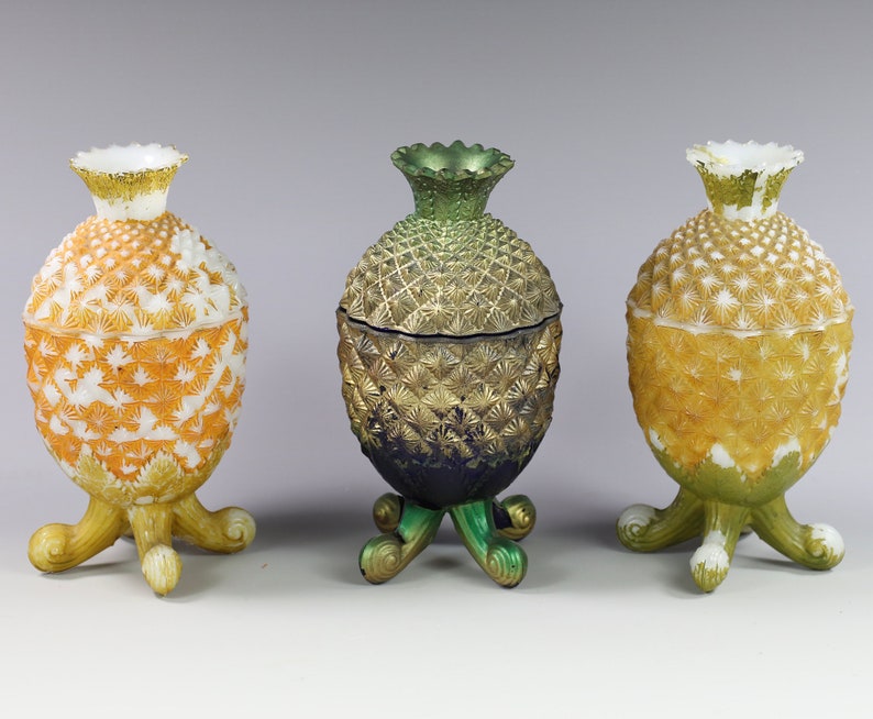 May include: Three decorative pineapple-shaped containers with lids. The containers are made of glass and have a white, yellow, and green color scheme. The containers are on a white background.