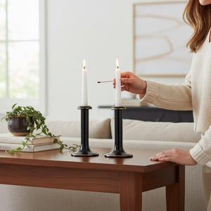 Puede incluir: Dos candelabros negros con bordes dorados, cada uno con una vela cónica blanca, alta y delgada. Las velas están colocadas en posición vertical sobre una superficie de madera, y una de ellas se enciende con una cerilla cerca de la mecha.