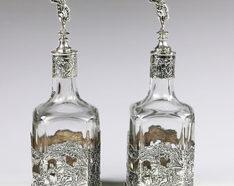 May include: Two silver decanters with ornate, detailed designs. Each decanter has a silver stopper with a small, silver figure on top. The decanters are clear glass and have a square shape.
