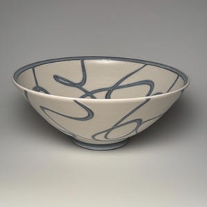 May include: A ceramic bowl with a smooth off-white matte finish featuring dark blue hand-painted swirling lines that wrap around the exterior and interior. The bowl has a wide circular rim and a small circular base, both accented in dark blue.