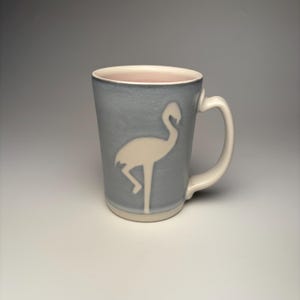 Op de afbeelding: Een keramische mok met een lichtblauwe buitenkant en een wit flamingo silhouet. De mok heeft een witte handgreep en een lichtroze binnenkant. De mok is een standaard maat.