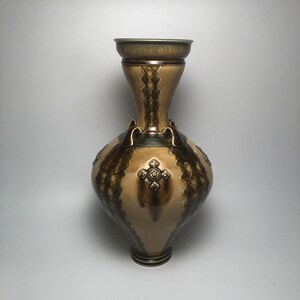 May include: A tall, ceramic vase with a wide, round body and a narrow neck. The vase is decorated with a brown and green geometric pattern and has a raised, cross-shaped design on the front.