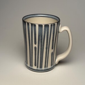 May include: A ceramic mug with a cream-colored base and a series of vertical, dark gray, brushstroke-style stripes. The mug features a smooth, curved handle on the right side and a dark gray rim that matches the vertical stripe pattern on the exterior.