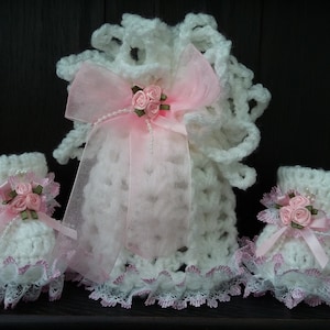 Op de afbeelding: Een set van drie gehaakte babyschoentjes met roze en witte kant en roze satijnen strikken. Elk schoentje heeft een trosje roze satijnen rozen.