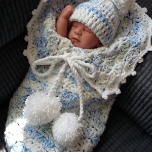 May include: A white and blue crocheted baby sleeping bag with a white pom-pom trim and a matching crocheted hat with a white pom-pom on top.
