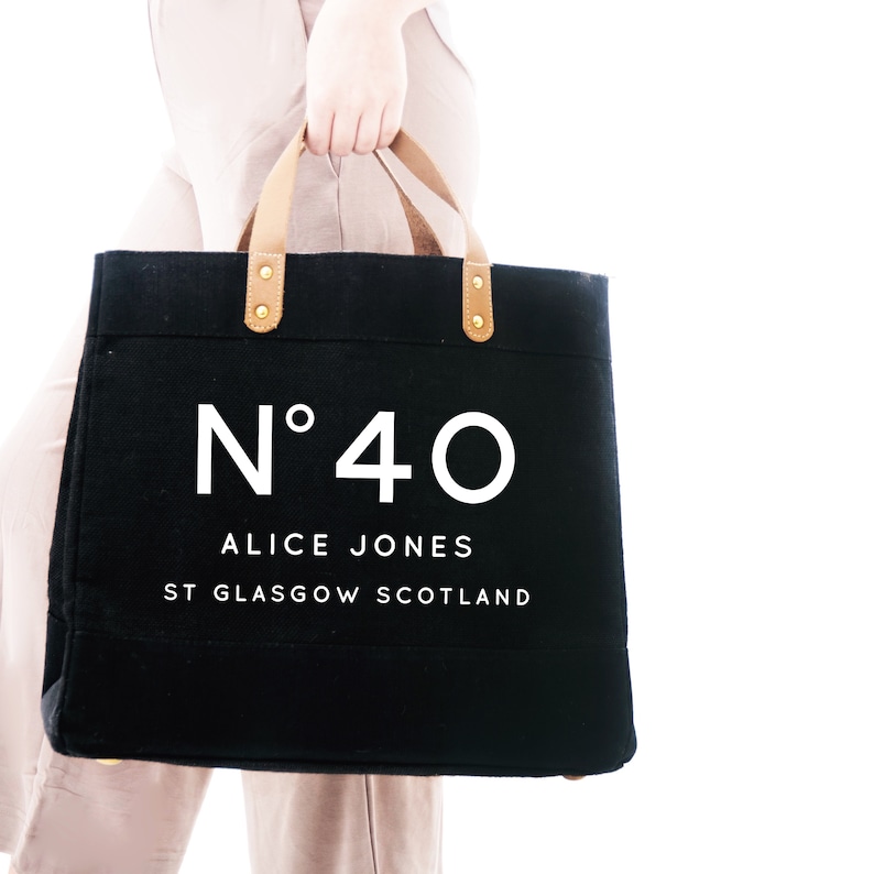 May include: A black canvas tote bag with brown leather handles and gold accents. The bag has the text "N&deg;40 ALICE JONES ST GLASGOW SCOTLAND" printed in white.