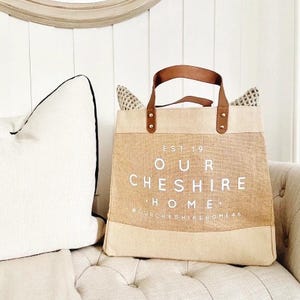 Puede incluir: Una bolsa de lona beige con asas de cuero marr&oacute;n y un logotipo blanco que dice "EST 19 OUR CHESHIRE HOME" con el texto "OURCHESHIREHOME46" debajo.