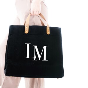 Puede incluir: Una bolsa de lona negra con asas de cuero marr&oacute;n y detalles dorados. La bolsa tiene las iniciales "LM" impresas en blanco en la parte delantera.