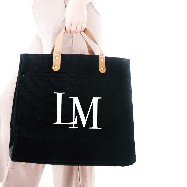 Puede incluir: Una bolsa de lona negra con asas de cuero marr&oacute;n y detalles dorados. La bolsa tiene las iniciales "LM" impresas en blanco en la parte delantera.