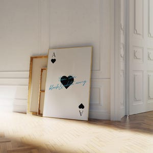 May include: A large white framed art print leaning against a wall, featuring a black heart and the text "toon town black & white army". The print is designed to resemble an Ace of Spades playing card. Two other framed canvases are behind it.