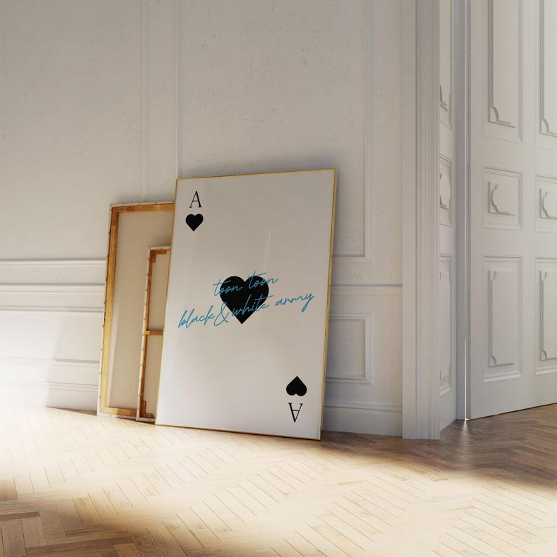 May include: A large white framed art print leaning against a wall, featuring a black heart and the text "toon town black & white army". The print is designed to resemble an Ace of Spades playing card. Two other framed canvases are behind it.