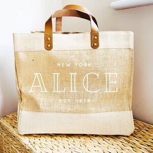 May include: A beige and brown tote bag with a white leather handle and gold accents. The bag has the text "NEW YORK ALICE EST 1979" printed on the front.