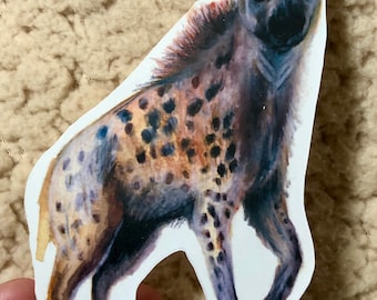May include: A watercolor illustration of a spotted hyena standing on all fours. The hyena is facing the viewer with its head tilted slightly to the side. The hyena is painted in shades of brown, black, and white.
