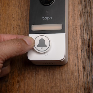Puede incluir: Un timbre blanco con una lente de cámara negra y la palabra "tapo" impresa en él. Una mano está presionando el botón del timbre, que tiene un icono de campana gris.