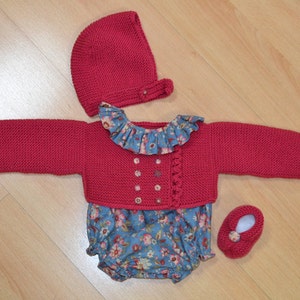May include: A red knitted baby outfit with a matching hat and booties. The outfit features a ruffled collar and a floral print on the bodice.