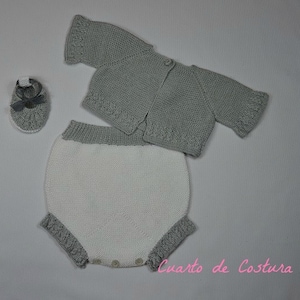 Puede incluir: Un conjunto de ropa de bebé tejido en gris y blanco, que incluye un cárdigan, bombachos y un par de zapatos. El conjunto está colocado sobre una superficie gris.